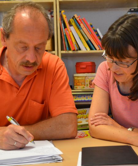 Der Leiter der Ambulanten Hilfen Untermain sitzt zusammen mit einer Kollegin am Schreibtisch. Beide beugen sich über einen Stapel Papiere, der die aktuellen Maßnahmen zur Hilfe für ein betreutes Kind bzw. einen betreuten Jugendlichen beinhaltet. Im Hintergrund ist ein Regal mit verschiedenen Sach- und Kinderbüchern.