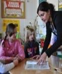 Drei Kinder sitzen an einem Tisch in einem Klassenzimmer. Im Hintergrund sieht man eine Tafel und Projektplakate und bunte Bilder an der Wand. Eine Frau steht bei den Kindern. Sie beugt sich zu den Papieren auf dem Tisch und erklärt etwas. Die Kinder schauen Aufmerksam zu.